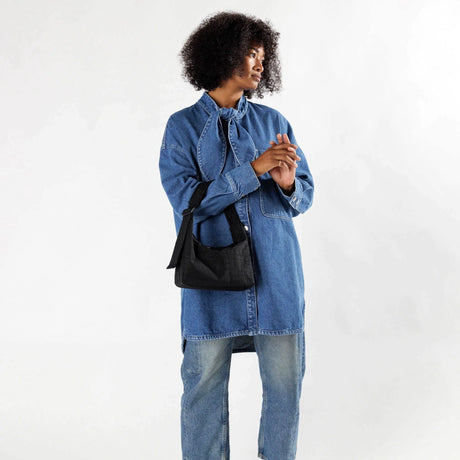 A person with curly hair wears a long denim shirt, light blue jeans, and carries the Baggu Mini Nylon Shoulder Bag in Black by Baggu while standing against a plain white background.