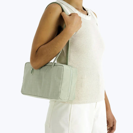 Muted sage green rectangular shoulder bag with top zipper and double handles worn over shoulder on person in cream outfit