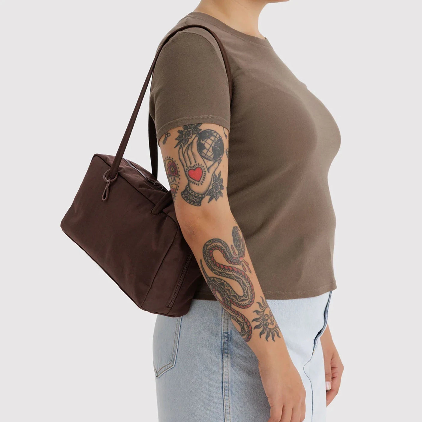 Dark brown rectangular shoulder bag with slim handles worn by tattooed person in brown t-shirt and light blue jeans