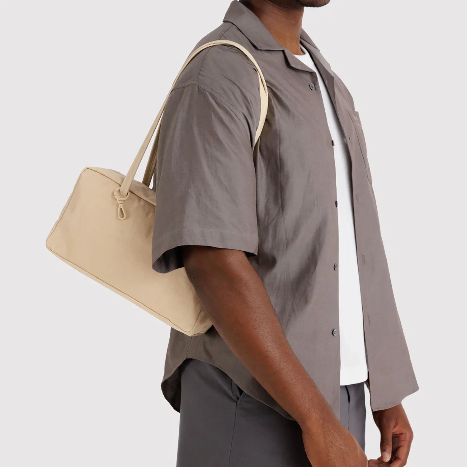 Beige rectangular fabric shoulder bag with slim double handles worn over shoulder by person in gray shirt and pants