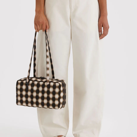 Rectangular shoulder bag with black and cream optical illusion pattern and long straps held by person in white pants
