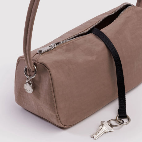 Rectangular muted taupe handbag with silver zipper and round metal charm attached to strap on plain background