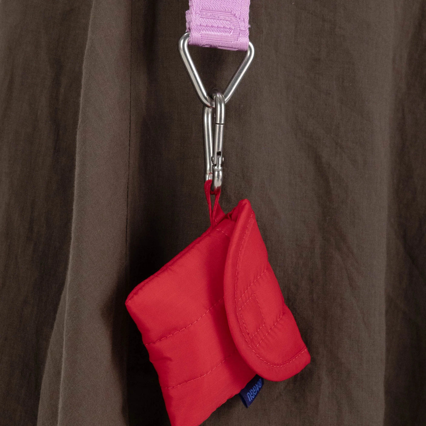 The Baggu Puffy Earbuds Case in Candy Red, ideal for AirPods Pro 2, is paired with a pink strap and silver carabiner against a brown fabric backdrop.