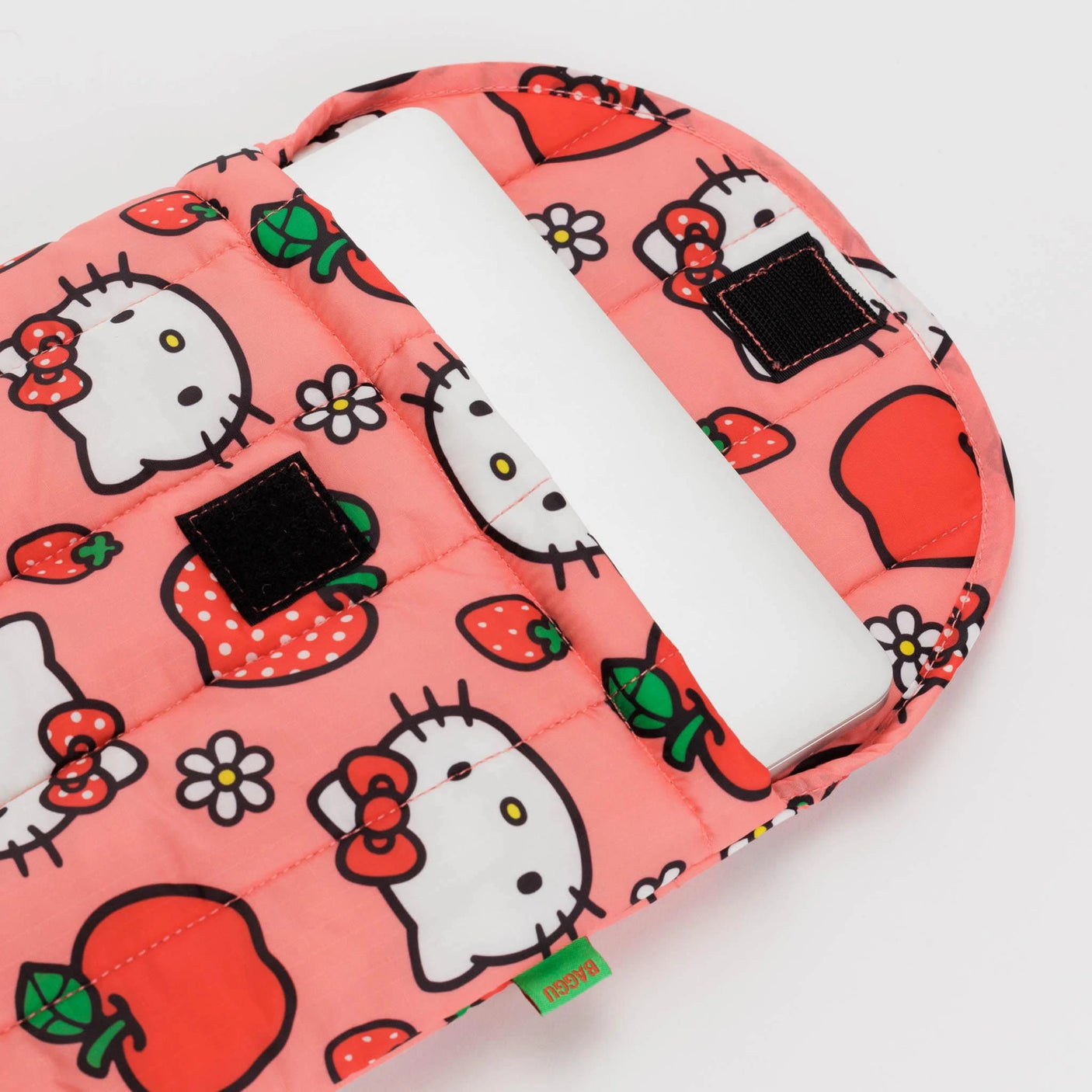 A pink Baggu laptop sleeve, part of the Hello Kitty collection, features a design with red apples and flowers. It's partially unzipped to reveal a white notebook and is crafted from recycled materials.