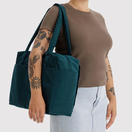 Dark teal fabric tote with wide shoulder straps and square silhouette carried by tattooed person in casual outfit
