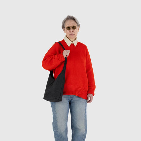 A person wearing sunglasses, a red sweater, and blue jeans carries a sleek black Baggu Small Nylon Sling - Black by Baggu on their shoulder against a plain background.