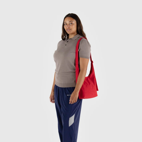 Person wearing a brown polo shirt, blue pants, and holding a Baggu Small Nylon Sling in Candy Apple, standing against a plain background.