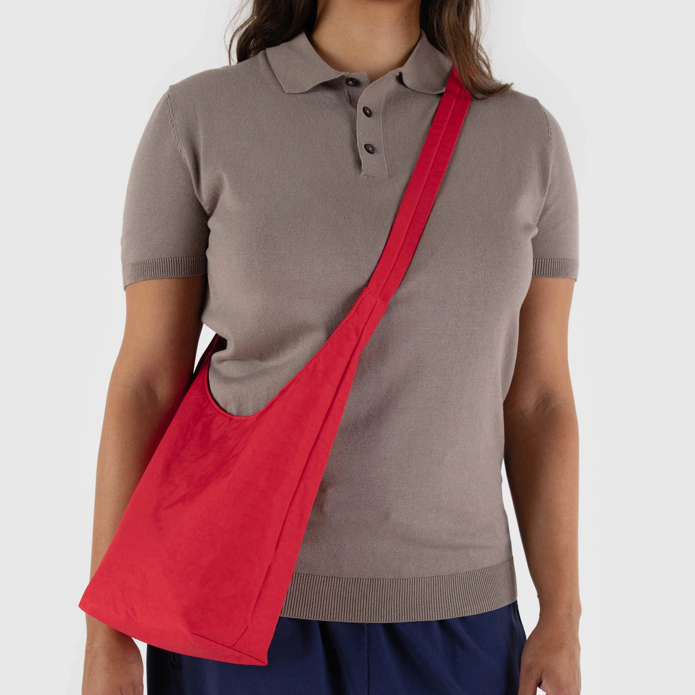 A person wearing a gray polo shirt and navy pants carries a Candy Apple Baggu Small Nylon Sling.