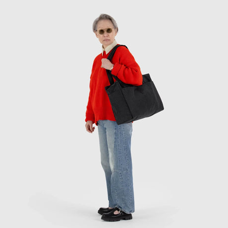 An older adult in a red sweater and sunglasses confidently stands against a white backdrop with the Baggu Small Cloud Carry-on - Black, a sleek tote made from recycled heavyweight nylon. Their short gray hair complements their blue jeans and black shoes, blending style and sustainability effortlessly.
