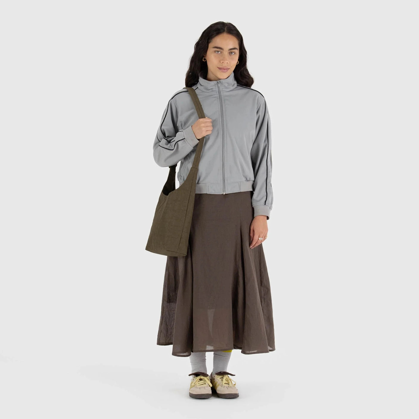 A person in a gray jacket and brown skirt stands holding a Baggu Small Nylon Sling - Seaweed, noted for its sustainable style and adjustable strap.