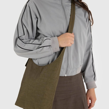 A person in a gray jacket carries a large Baggu Small Nylon Sling in Seaweed, featuring an adjustable strap and highlighting their sustainable style.