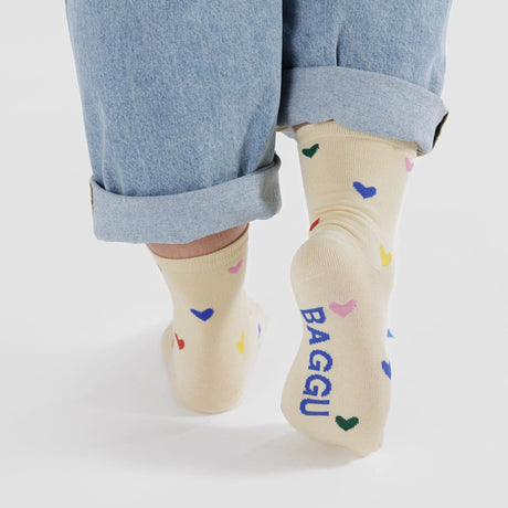 A person is shown from the back wearing rolled-up jeans and Baggu Crew Socks - Hearts, which are cream-colored with embroidered multicolored hearts and feature "BAGGU" on one sock.