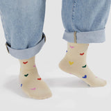 A person wearing rolled-up jeans and cream-colored Baggu Crew Socks, featuring embroidered multicolored heart patterns.