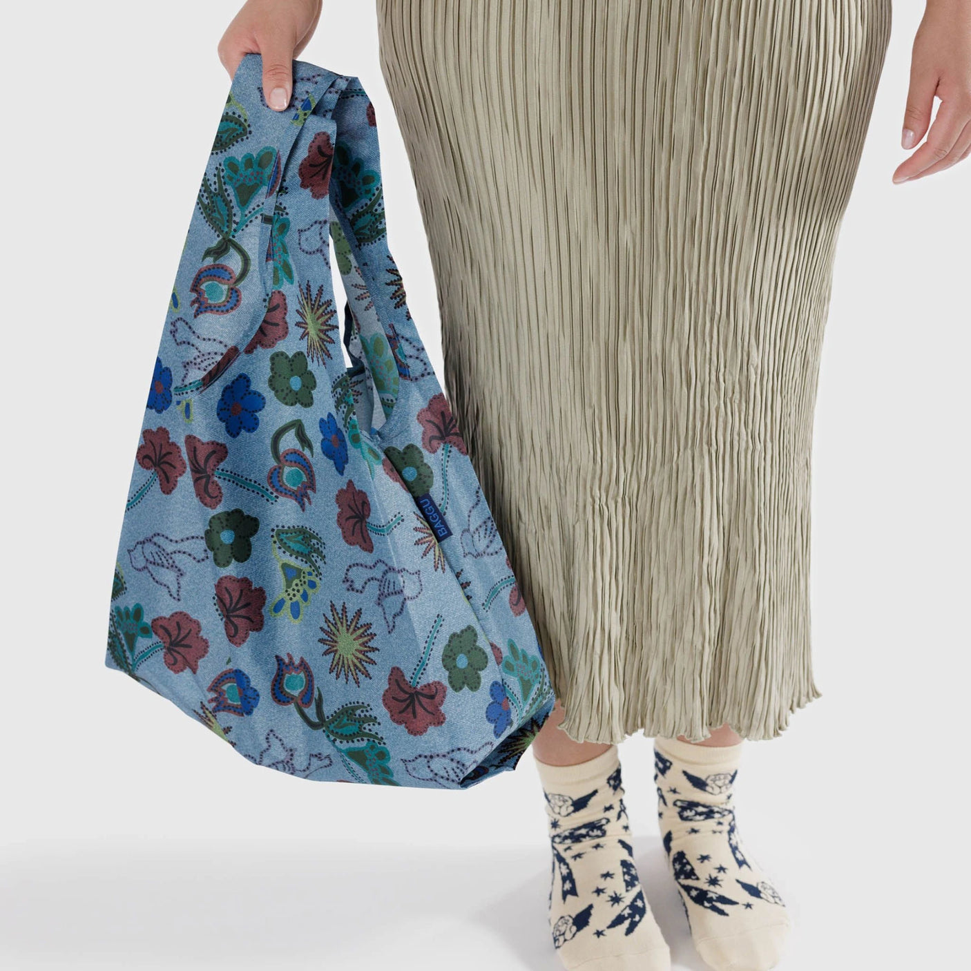 A person holds a Baggu Standard Reusable Bag in the Digital Denim Birds design while showcasing their style in a chic pleated skirt and patterned socks.