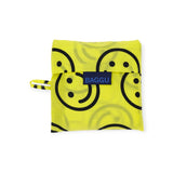 A folded Baggu Standard Reusable Bag - Yellow Happy rests on a white background, showcasing a smiley face design and a blue label. This eco-friendly tote from Baggu combines style and sustainability, making it the perfect reusable bag for your everyday needs.