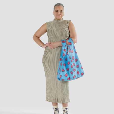 A person wearing a sleeveless beige dress is holding the Baggu Standard Bag - Keith Haring Hearts, a vibrant blue reusable bag with red heart patterns, while standing against a plain white background.