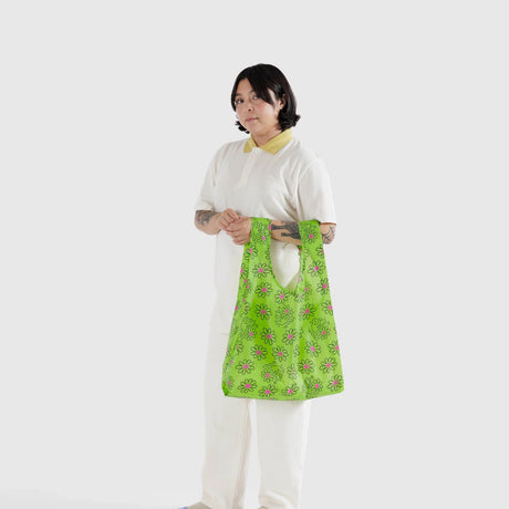 A person dressed in white holds the Baggu Standard Bag - Keith Haring Flower, featuring a green floral pattern and made from recycled nylon yarn, against a plain background.