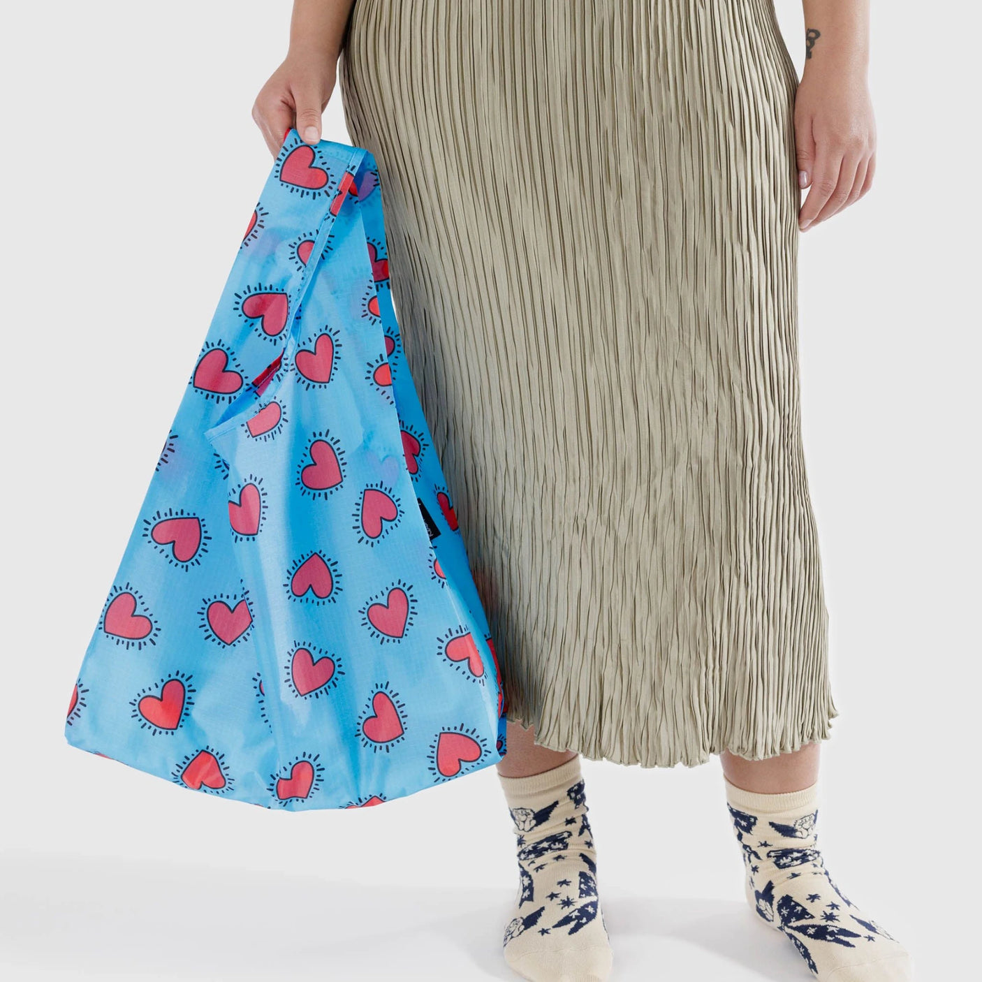 A person is holding a Keith Haring x Baggu Standard Bag featuring red heart designs, while wearing a pleated skirt and patterned socks.