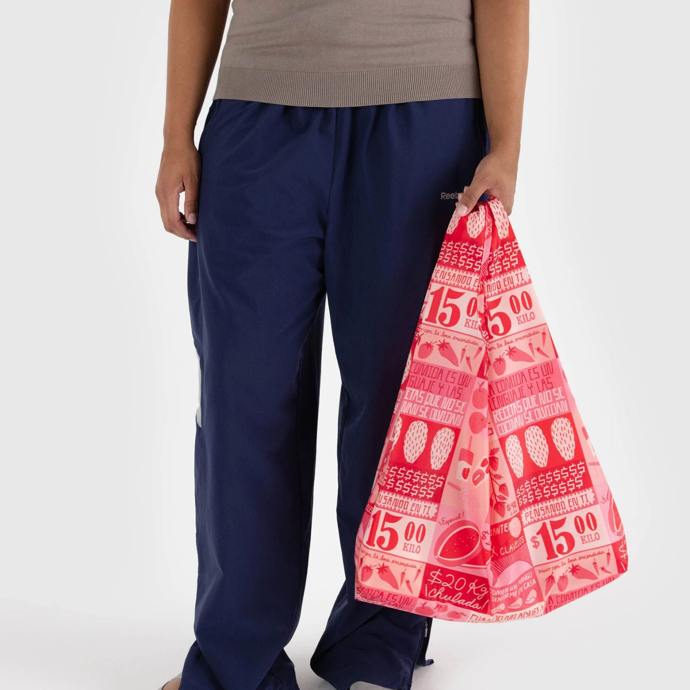 A person is wearing a beige top and blue pants while holding the eco-friendly Baggu Standard Reusable Bag - Mercado from Baggu, which showcases a lively design of numbers and strawberries made from recycled nylon.