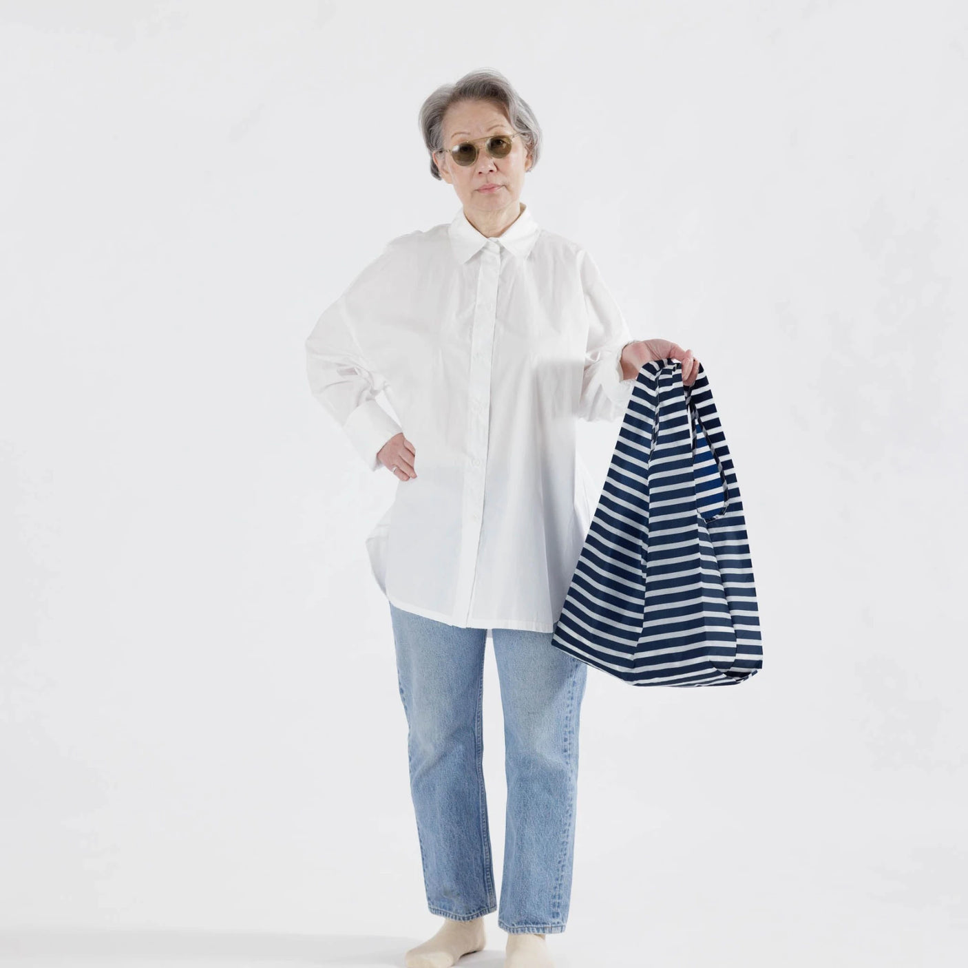 An older adult wearing a white shirt, blue jeans, and sunglasses holds a Baggu Standard Reusable Bag in Navy Stripe.