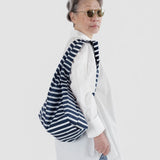 A person with sunglasses and a white shirt holds the Baggu Standard Reusable Bag in Navy Stripe, crafted from recycled nylon, over their shoulder.