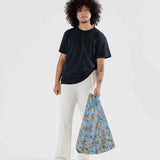 A person with curly hair, dressed in a black shirt and white pants, is holding a Baggu Standard Reusable Bag - Orchid, made from recycled nylon.