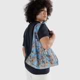 A person with curly hair is dressed in a black t-shirt and white pants, holding a large Baggu Standard Reusable Bag - Orchid, made of recycled nylon with a blue floral design, over their shoulder against a plain background.