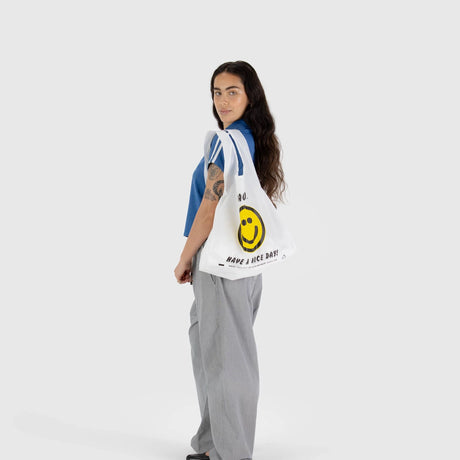 A woman with long hair is wearing a blue shirt and gray pants while holding the Baggu Standard Reusable Bag in the "Thank You Happy" design. This bag, from the brand Baggu, features a smiley face and the phrase "Have a Nice Day!" printed on it. It's an eco-friendly accessory made from recycled nylon and offers large capacity storage, combining everyday charm with sustainable design.