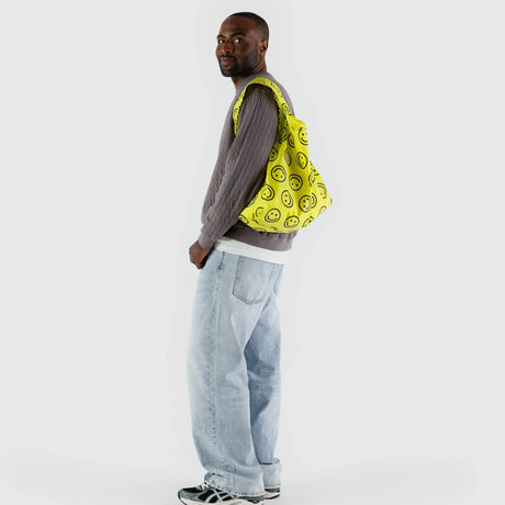 A man wearing a gray sweater and light jeans stands against a plain white background, carrying the Baggu Standard Reusable Bag - Yellow Happy, featuring a smiley face design.