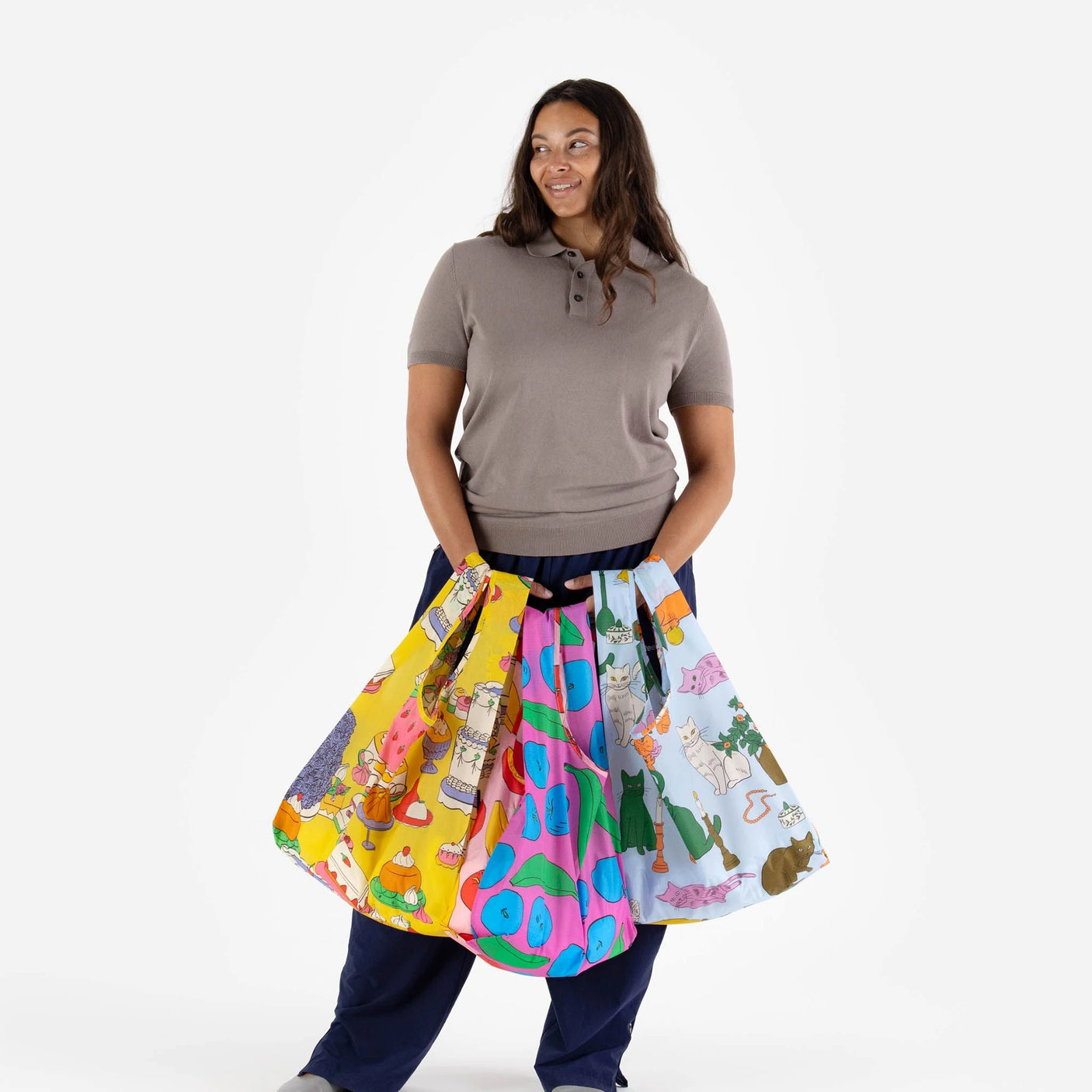 A person in a gray shirt holds the Baggu Set of 3 Standard Bags - Still Life, crafted from recycled nylon, featuring three colorful bags with various patterns, standing against a plain white background.