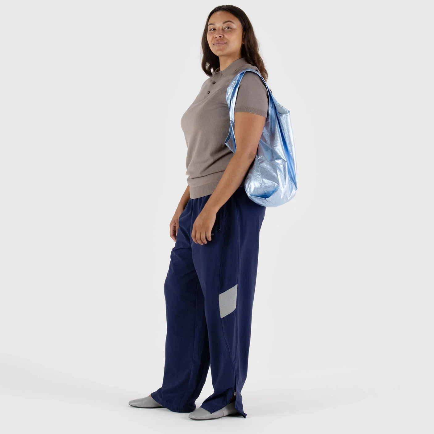 A woman stands against a white background, wearing a beige top and blue pants. Over her shoulder is the Baggu Standard Reusable Bag in Metallic Light Blue, crafted from recycled nylon by Baggu, adding an eco-friendly flair to her stylish ensemble.