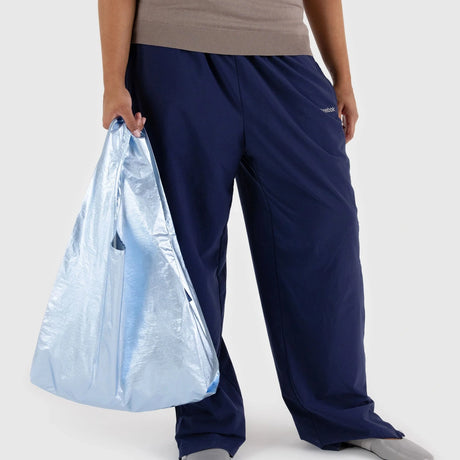 A person wearing dark blue pants and a beige top holds a metallic light blue reusable bag from Baggu, crafted from recycled nylon.