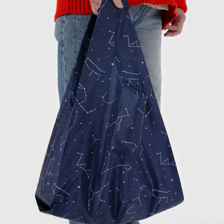 A person holding a Baggu Standard Reusable Bag - Constellation, crafted from recycled nylon in navy blue and featuring white constellation patterns.