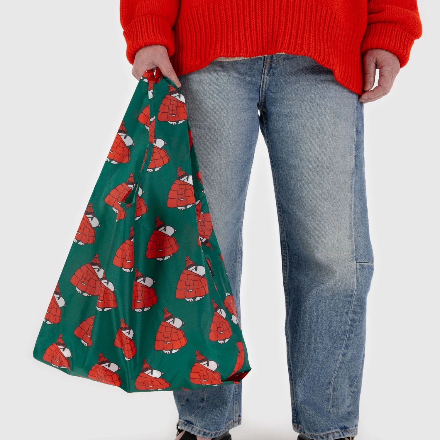 The individual wearing blue jeans and a red sweater is holding a Baggu Standard Reusable Bag - Puffer Snoopy Red, characterized by its green design adorned with a whimsical red turtle pattern.