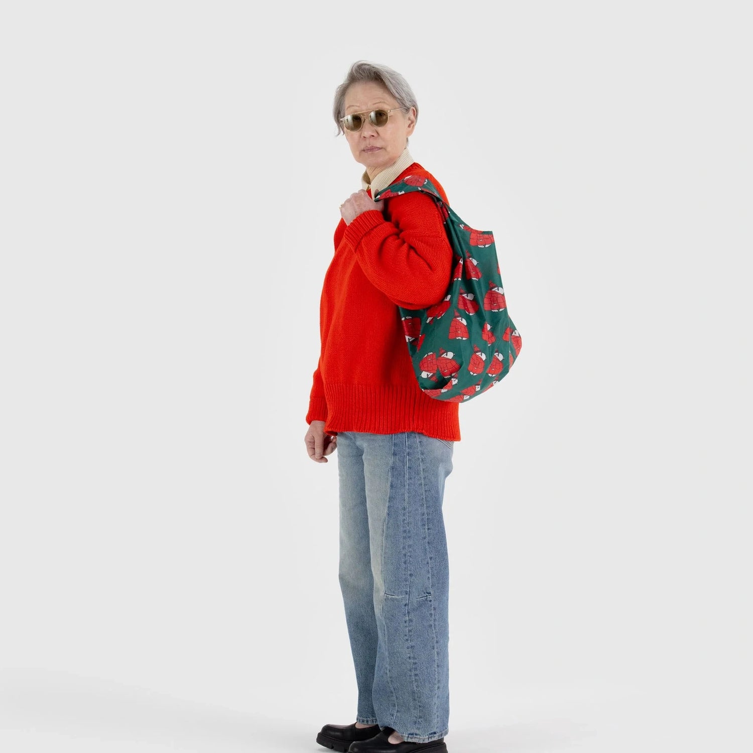 A person in a red sweater and jeans with sunglasses stands against a plain light background, carrying a Baggu Standard Reusable Bag - Puffer Snoopy Red.