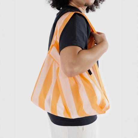 A person carries a Baggu Standard Reusable Bag - Tangerine Stripe on their shoulder, wearing a black shirt and light-colored pants, against a plain background.