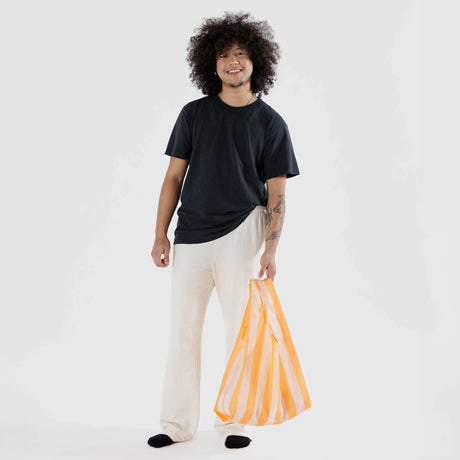 A person with curly hair, wearing a black t-shirt and light pants, holds a Baggu Standard Reusable Bag in Tangerine Stripe.