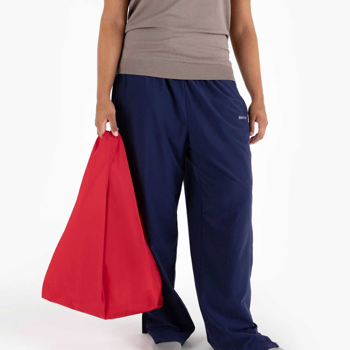 A person holding a Baggu Standard Reusable Bag in Candy Apple, wearing a taupe shirt and navy blue pants, stands against a plain background.