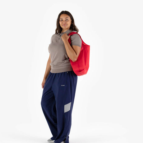 A person stands, holding a Baggu Standard Reusable Bag in candy apple red. They are wearing a grey polo shirt and blue pants.