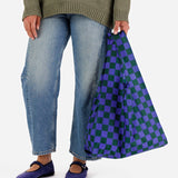 A person wearing blue jeans and a green sweater is holding a stylish Baggu Standard Reusable Bag - Iris Green Check, featuring a vibrant green and blue checkered pattern, made from durable recycled nylon.