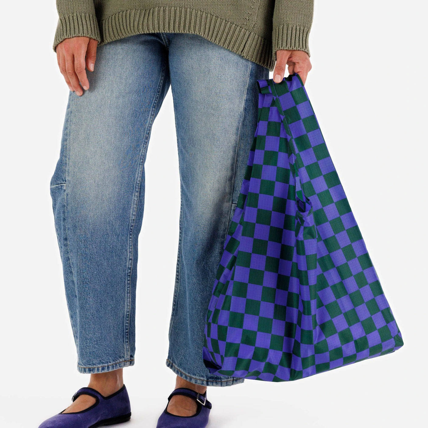 A person in blue jeans and a green sweater is holding a jewel-toned Baggu Standard Reusable Bag from the Baggu Set of 3 Standard Bags - Jewel Checks, made of recycled nylon. They are wearing purple shoes.