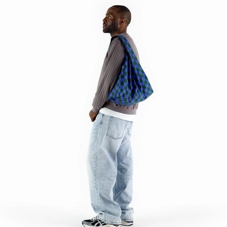 A person in a gray sweater and loose jeans stands sideways, carrying a Baggu Standard Reusable Bag in an iris green checkered pattern made from recycled nylon over their shoulder.