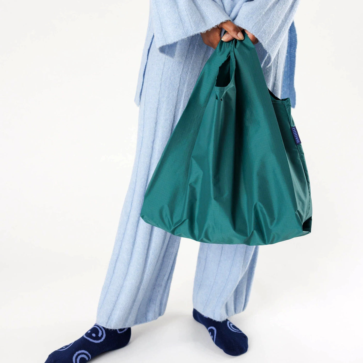 A person in light blue pants holds a Baggu Standard Reusable Bag in Malachite, made from recycled nylon.