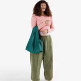 A person with curly hair in a pink long-sleeve shirt and green pants is standing against a plain background, holding the Baggu Standard Reusable Bag - Malachite, crafted from recycled nylon.