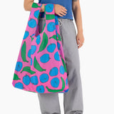 A person carries a Big Baggu Bag - Pink Apples & Bananas, an eco-friendly choice from Baggu, which perfectly complements their blue shirt and grey pants.