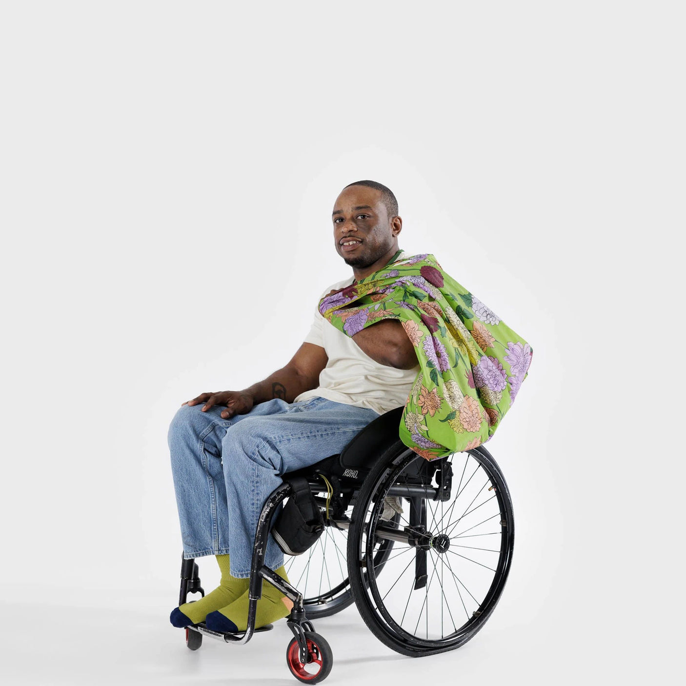 A man in a wheelchair poses with the Big Baggu Bag - Dahlia from the brand Baggu draped around his shoulders. He wears a light shirt, jeans, and yellow socks with blue accents, set against a plain white background.