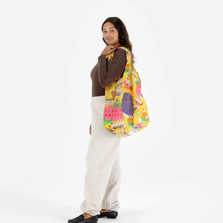 A person with long hair stands against a white backdrop, highlighting the eco-friendly Big Baggu Bag in the vibrant patisserie pattern from Baggu over their shoulder, paired with a brown top and light-colored pants.