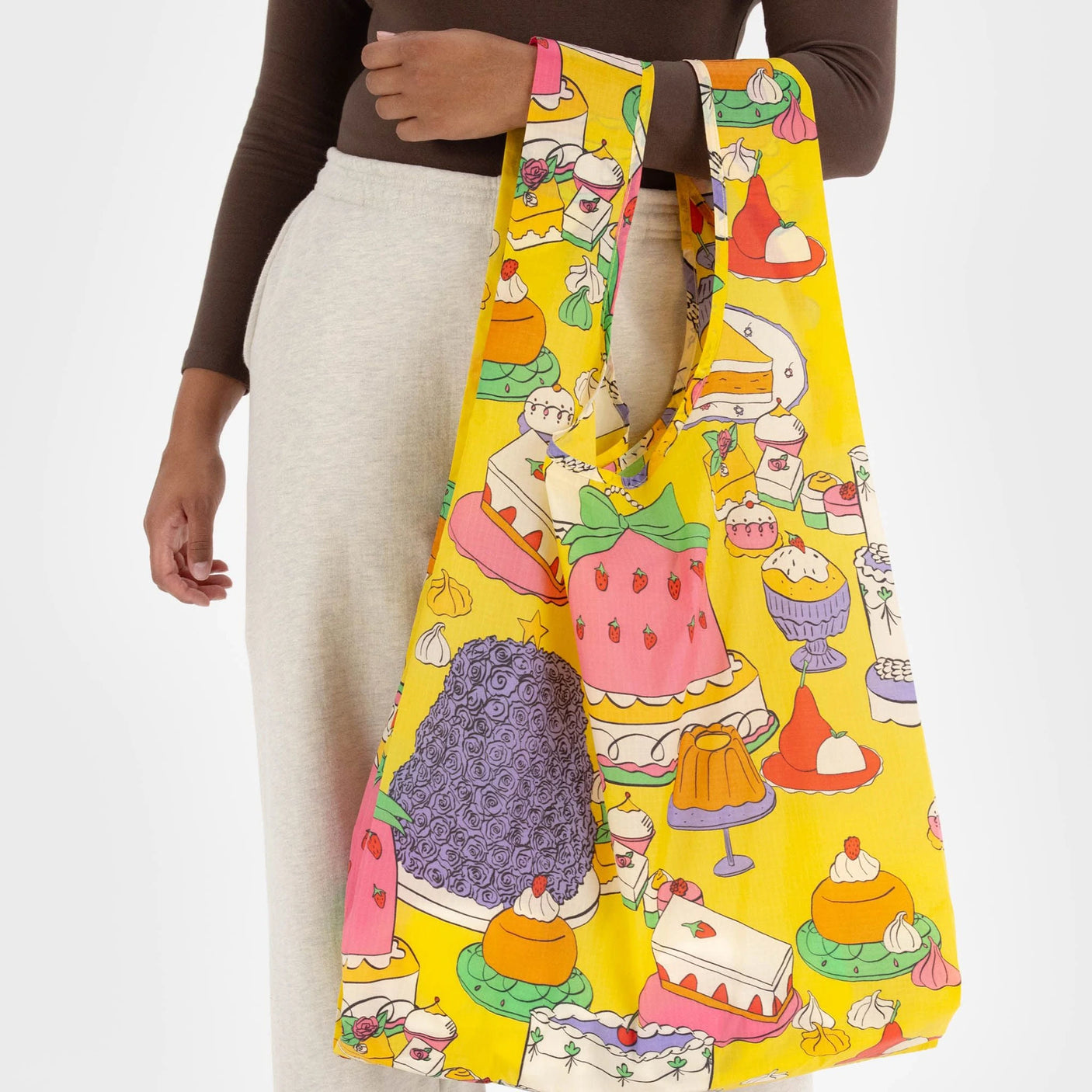 A person holds the eco-friendly Big Baggu Bag - Patisserie by Baggu, featuring a vibrant print of assorted cakes and desserts on a bright yellow backdrop.