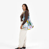 A person poses against a plain white background, holding the vibrant Big Baggu Bag in the whimsical "Table Cats" design by Baggu. They're dressed in a brown long-sleeve shirt and light-colored pants. This eco-friendly tote features recycled nylon, adding sustainability to its colorful charm.