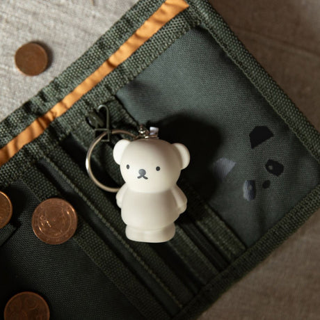Small white bear-shaped keychain with minimal facial features attached to green fabric wallet with coins nearby
